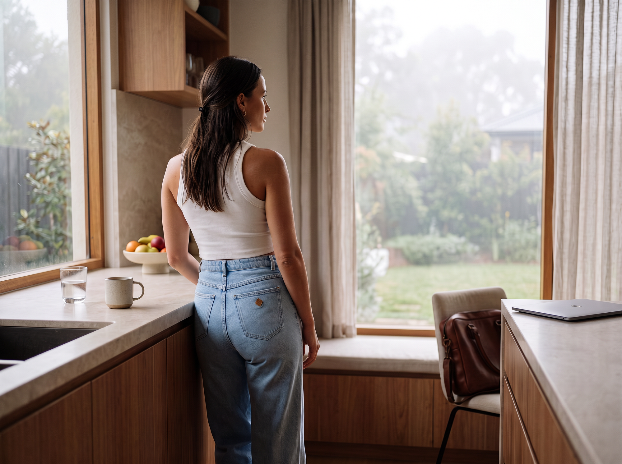Woman standing in a sunlit kitchen, looking out a window in quiet reflection. Warm morning light, natural timber and stone finishes, and everyday objects create a calm, thoughtful atmosphere.